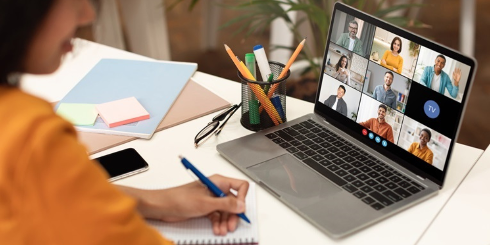 A woman sits at a desk with a notebook and pen with a laptop in front of her with Zoom on the screen.