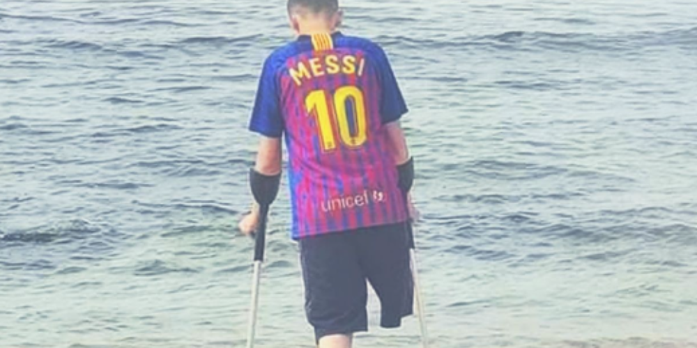 A young boy with one leg and using crutches stands at the edge of the ocean.