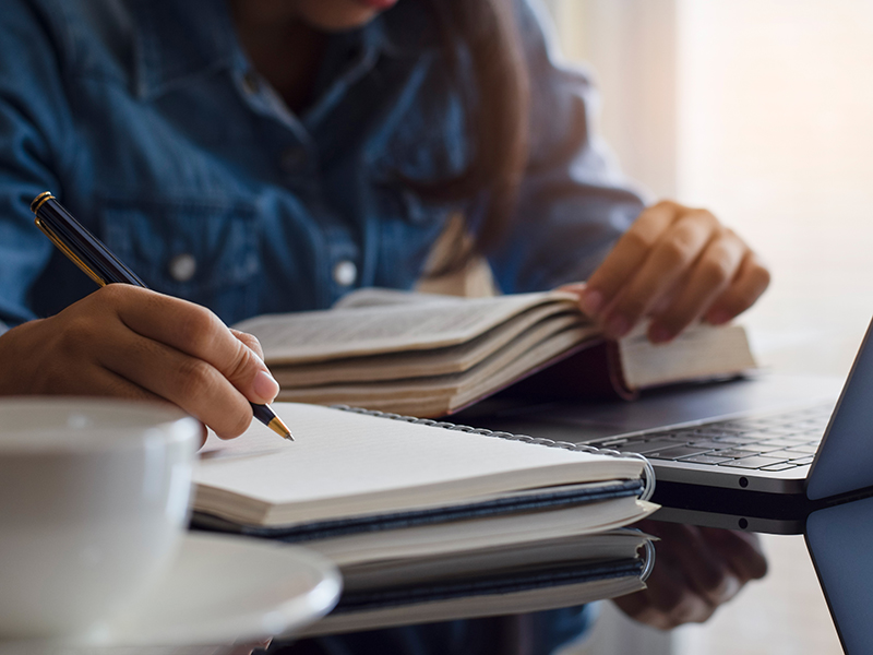 A learner writes notes in their notebook while they watch training on a laptop