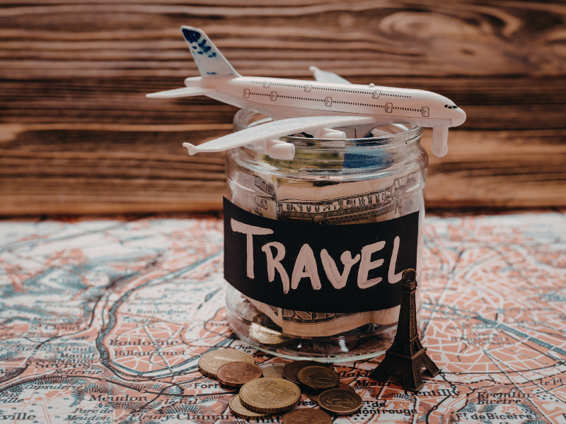 A small airplane sits on a Travel jar filled with money, sitting on a paper map