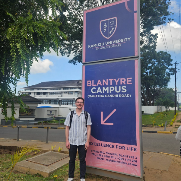 A student stands outside beside a sign for Kamuzu's Blantyre Campus